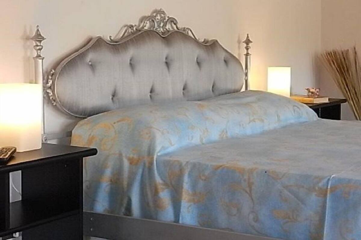 Ein Schlafzimmer mit einer gepolsterten Kopfstütze, einem Bett mit blauen Bettbezügen, einem Nachttisch mit Lampe sowie einem Beistelltisch mit Buch und einer Dekoration.