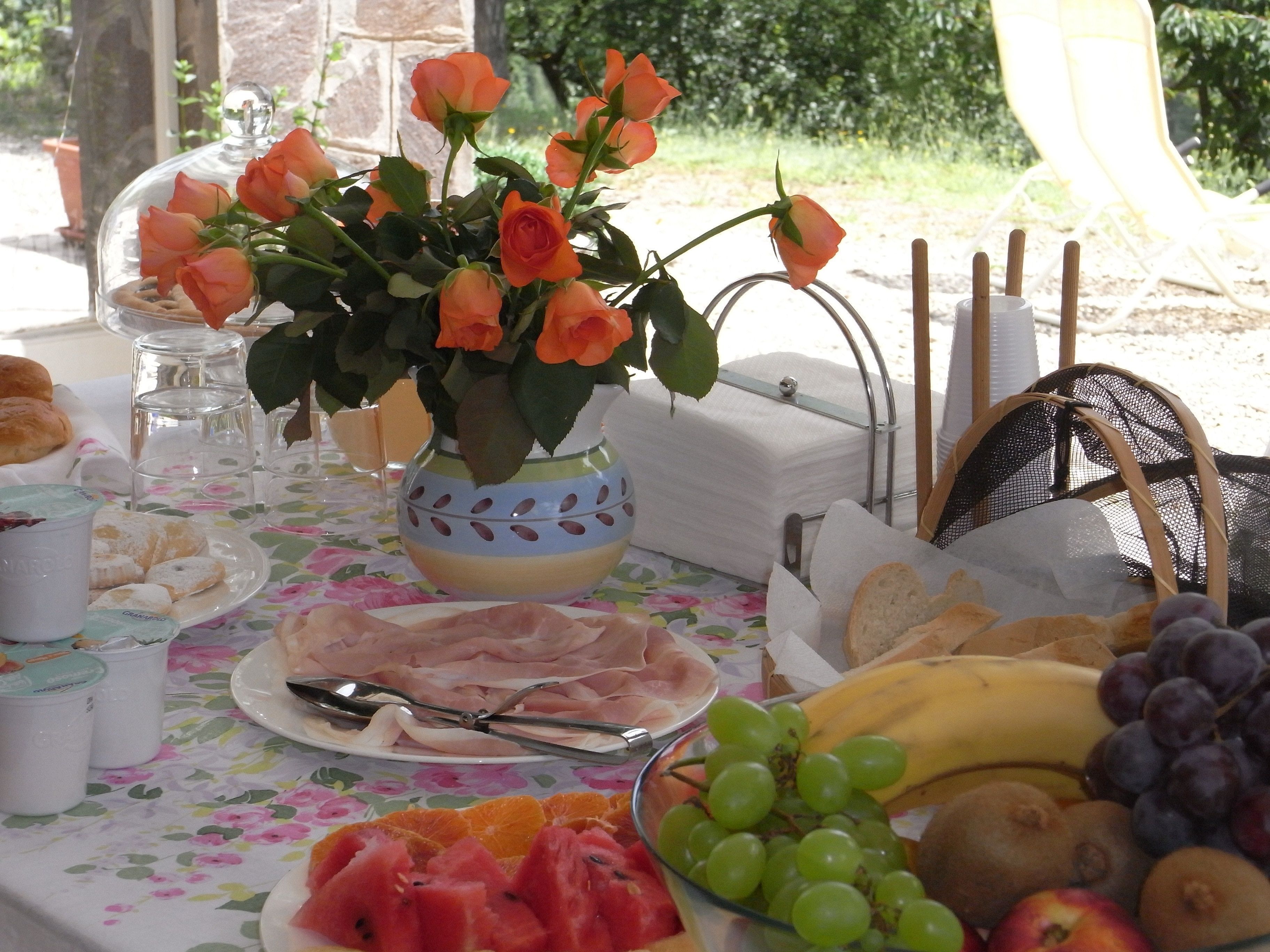 Ein Tisch reichhaltig gedeckt mit Speisen und Getränken, dazu eine Rosenvase, Tellervoll geschnittenem Obst und eine Schale Trauben.