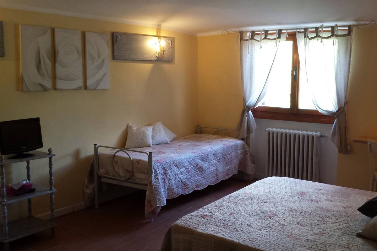 Ein Schlafzimmer mit einem Einzelbett, einem Sessel, einem Schreibtisch mit Computer und einem Heizkörper. Ein Fenster mit Vorhängen ist ebenfalls vorhanden.