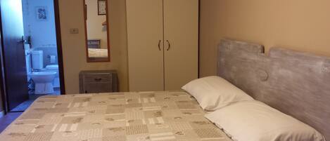 Un dormitorio con cama, mesita de noche, armario y baño a la vista a través de una puerta abierta.