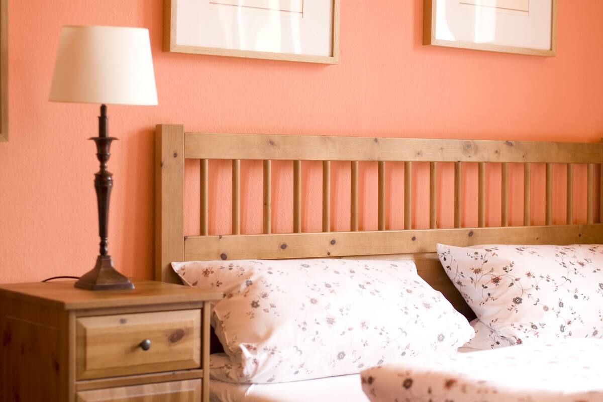 Un dormitorio con una cama de madera, una mesita de noche con una lámpara y dos impresiones botánicas enmarcadas en la pared.