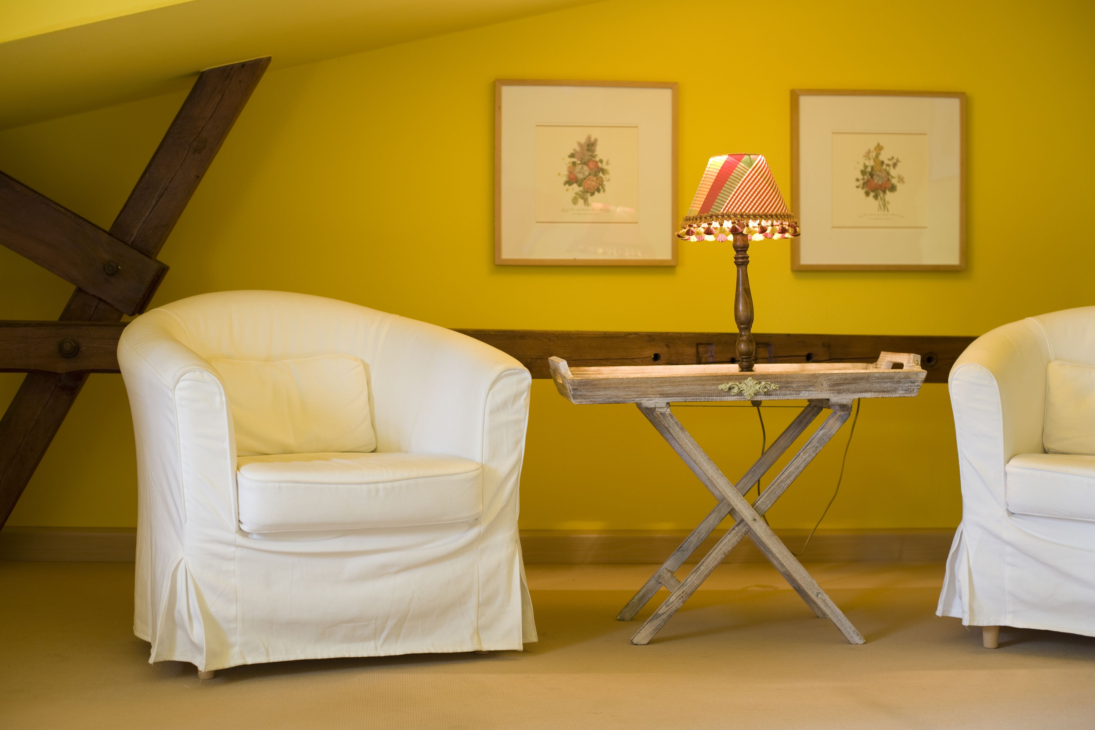 Una habitación con dos sillones blancos, una mesa de madera con una lámpara y dos cuadros con motivos florales en la pared amarilla.