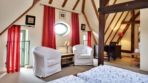 Een kamer met een schuine plafond, twee witte stoelen, een bed met bloemenbeddengoed, een bureau met een lamp en rode gordijnen.