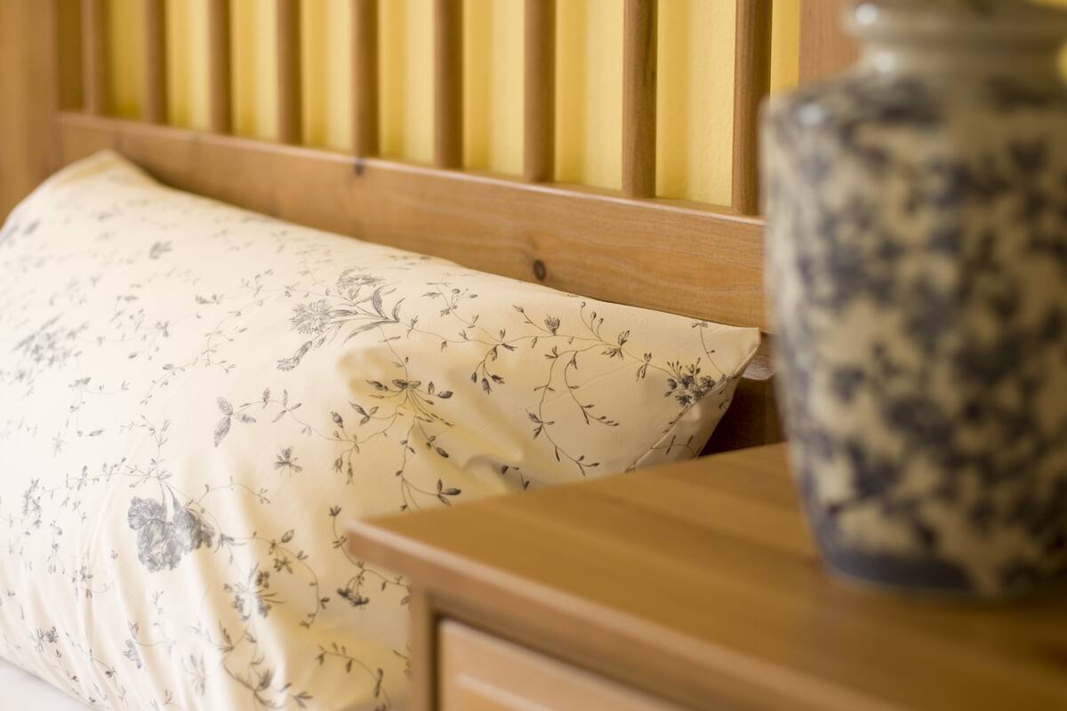 Una cama con un cojín estampado con motivos florales, un cabecero de madera y una mesita de noche con una lámpara azul y un jarrón de cerámica.