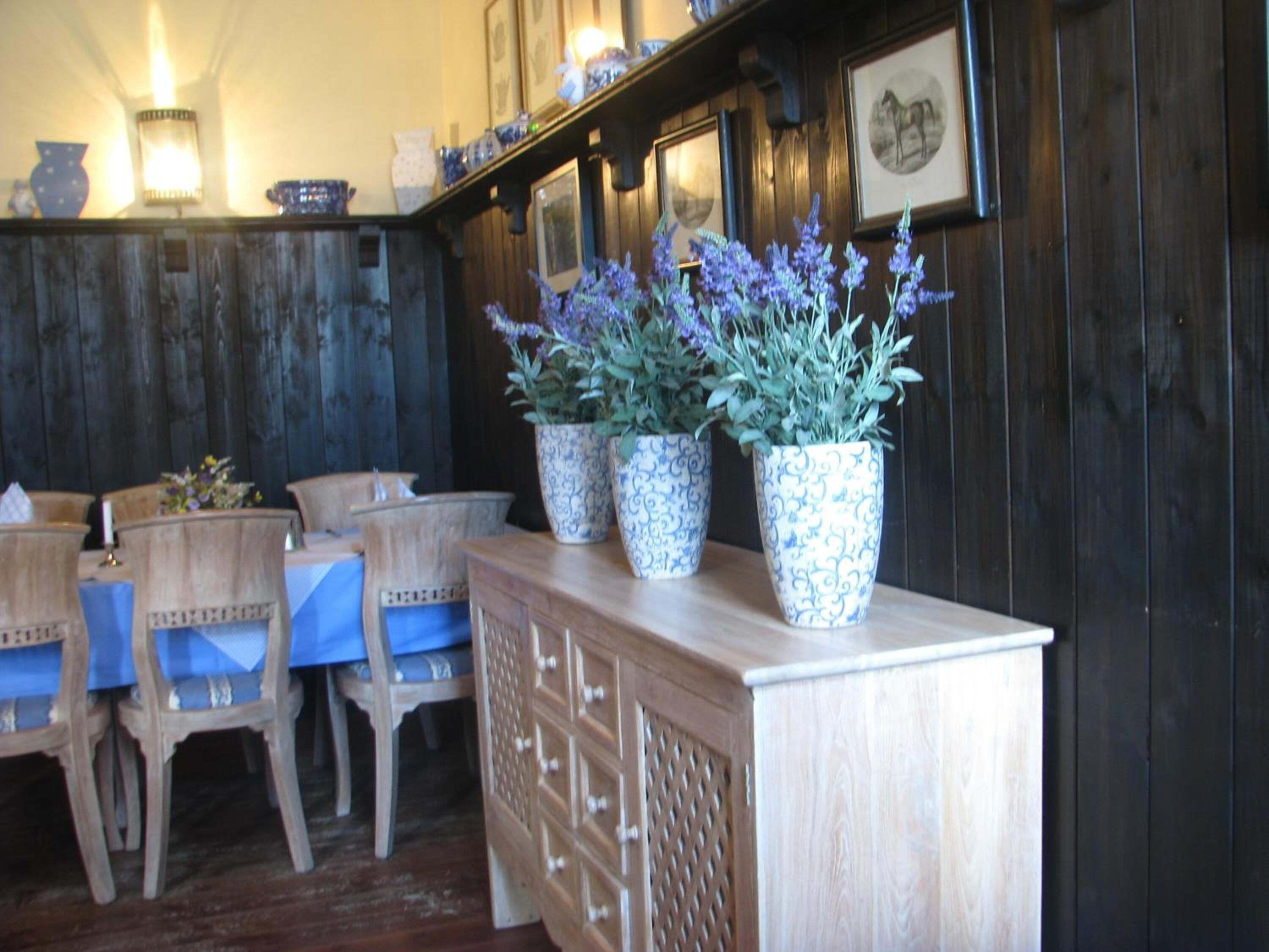 Een houten eethoek met blauwe stoelen, een houten buffet en decoratieve vazen met paarse bloemen.