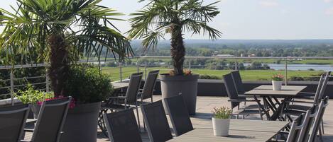 An outdoor terrace with tables and chairs, potted plants, and a view of a river and landscape.