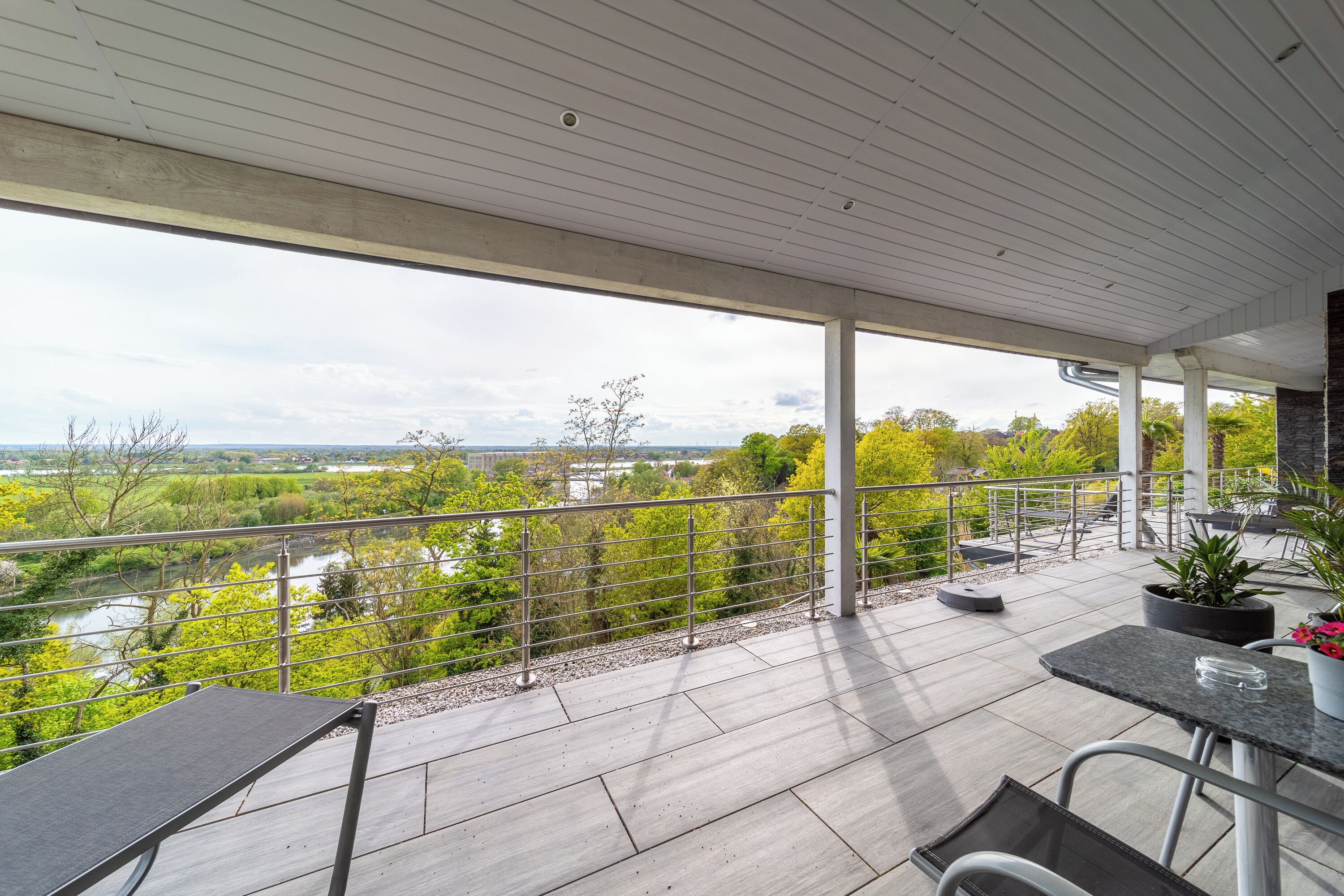 A covered outdoor area with a view of a river and greenery, featuring a table and chairs.