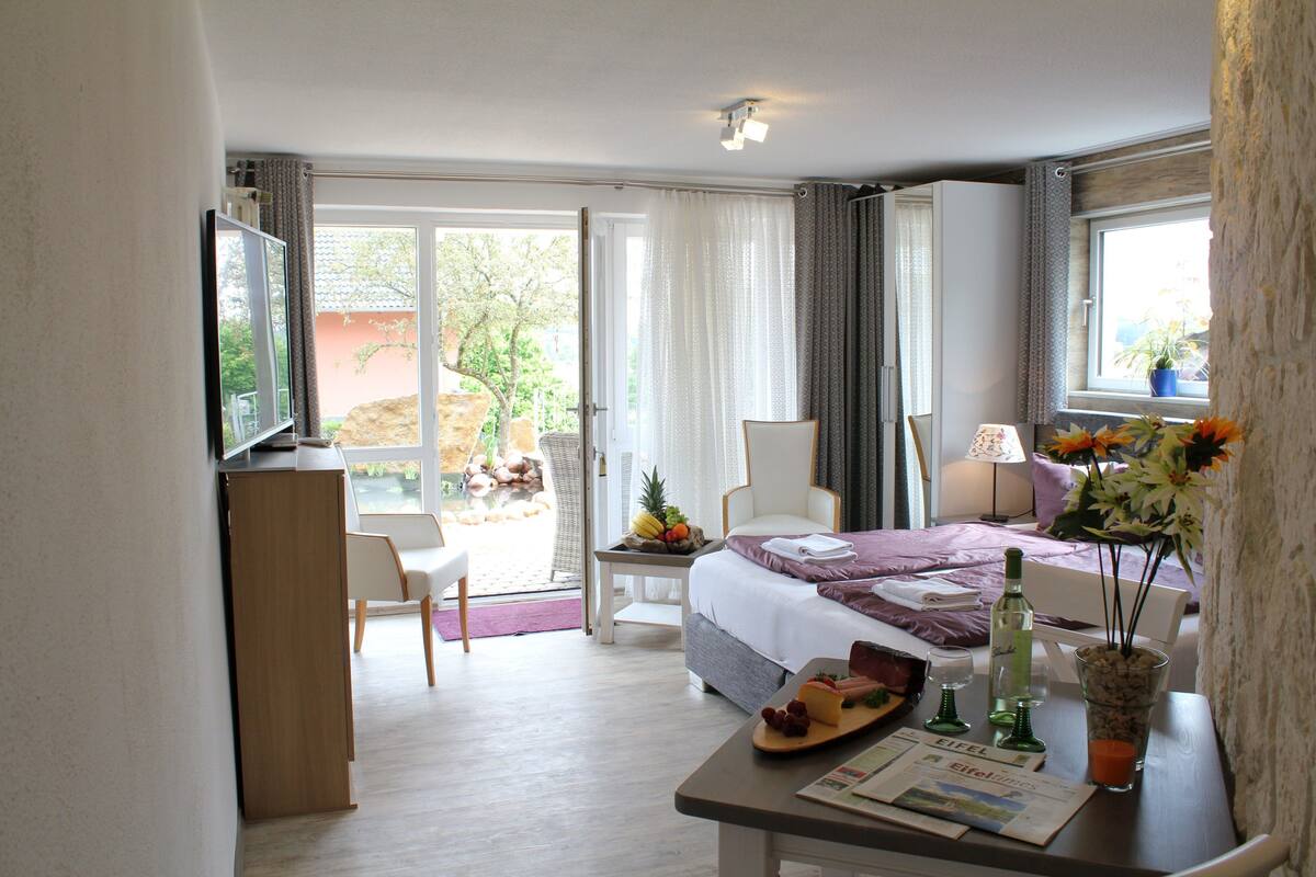 A hotel room with a bed, a desk with a laptop, a chair, a dining table with fruits and a bottle, and a window with a plant.