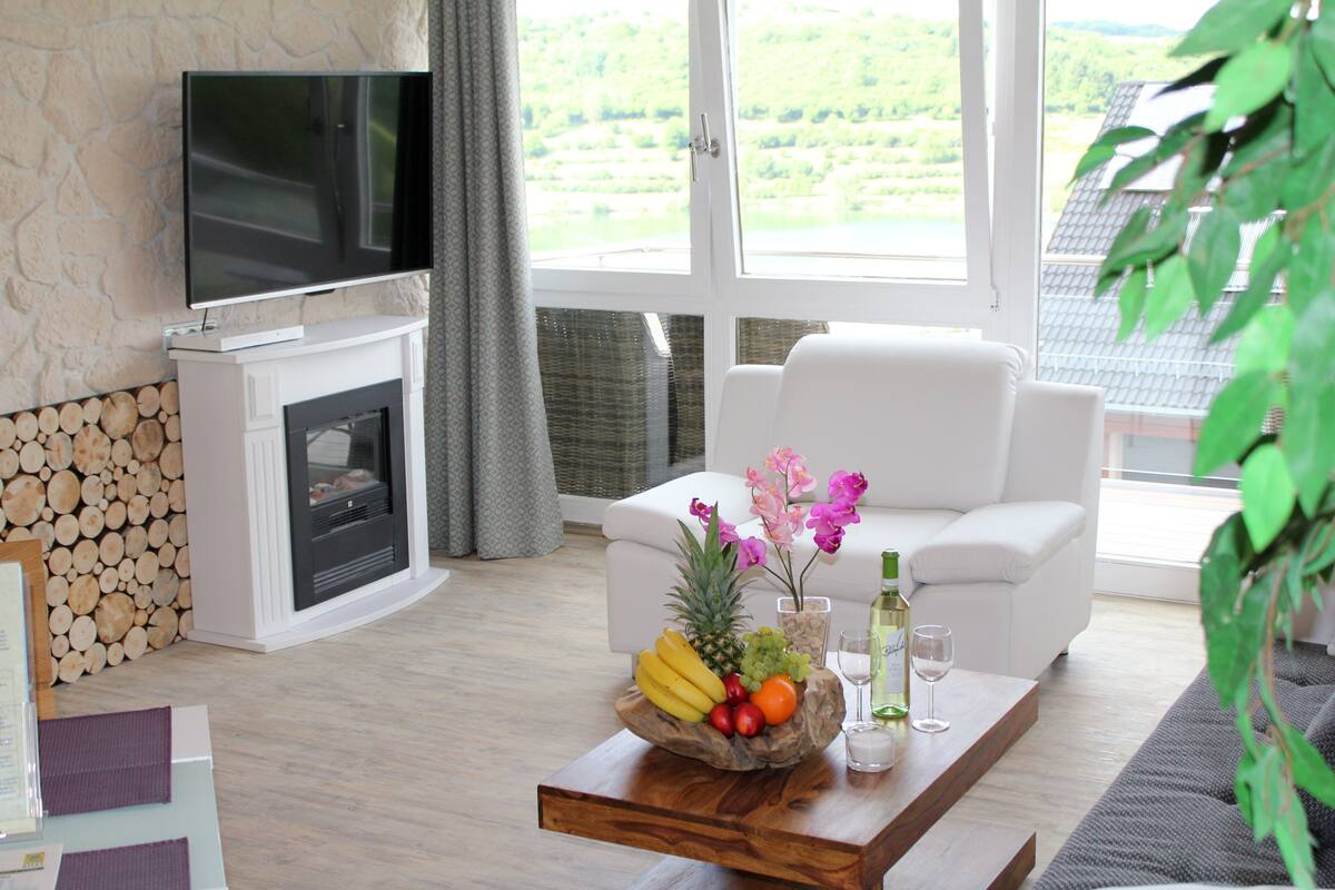 A modern living room with a fireplace, a flat-screen TV, a white sofa, and a coffee table with fruit and drinks.