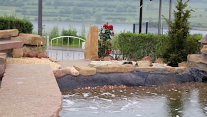 A stone pathway leading to a small pond with a metal railing and a flower arrangement. - Ferienwohnungen Maarberg Resort (Schalkenmehren)