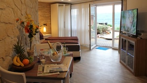 A hotel room with a bed, a television, a dining table with a fruit bowl, a newspaper, and a view of the outdoors. - Ferienwohnungen Maarberg Resort (Schalkenmehren)