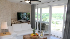 A modern living room with a white sofa, a wooden coffee table with fruit and a bottle, a flat-screen TV, stone wall, and a balcony with a scenic view. - Ferienwohnungen Maarberg Resort (Schalkenmehren)