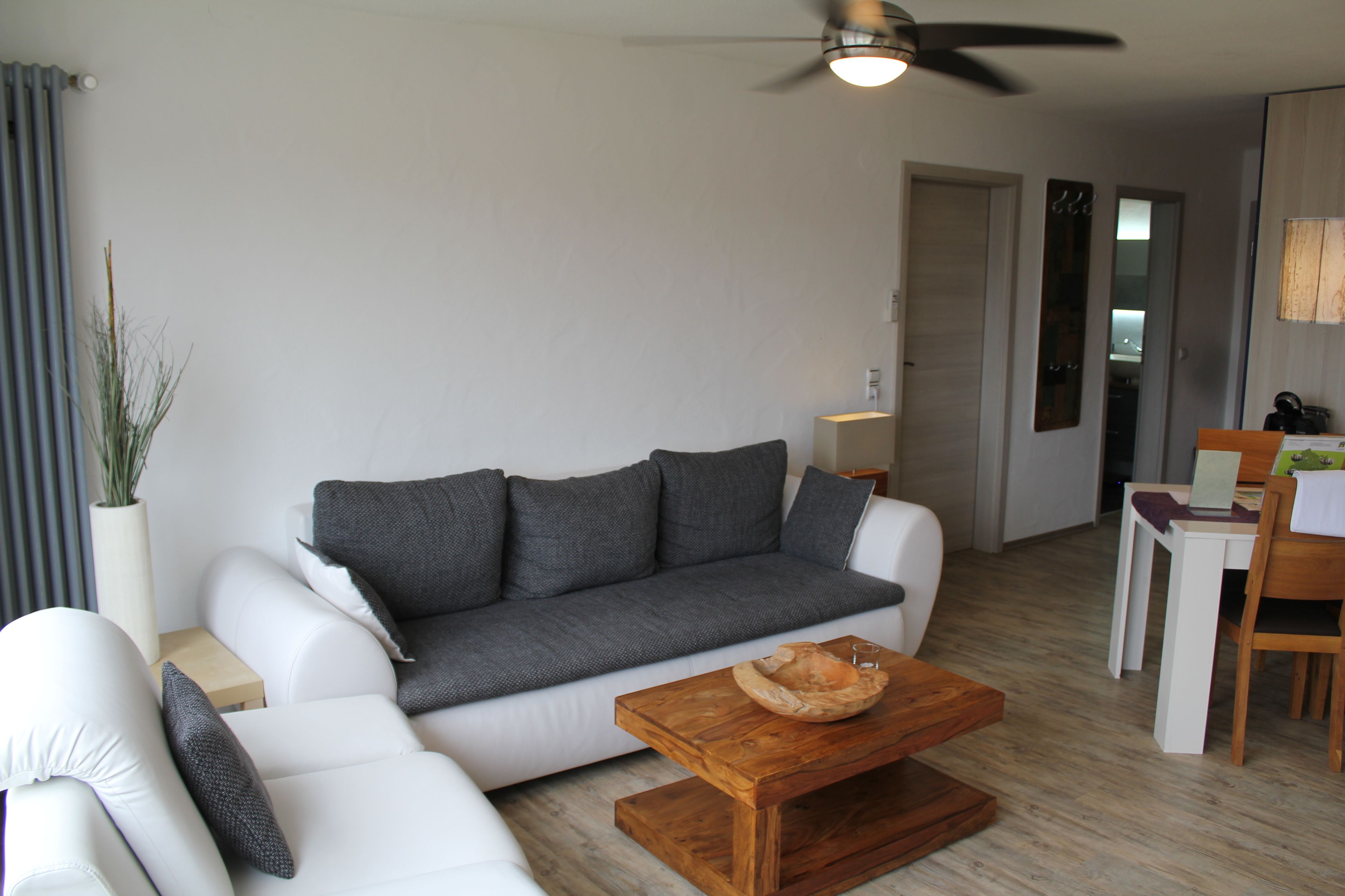 Ein modernes Wohnzimmer mit grauem Sofa, weißen Sesseln, einem Holztisch und einem Deckenventilator.
