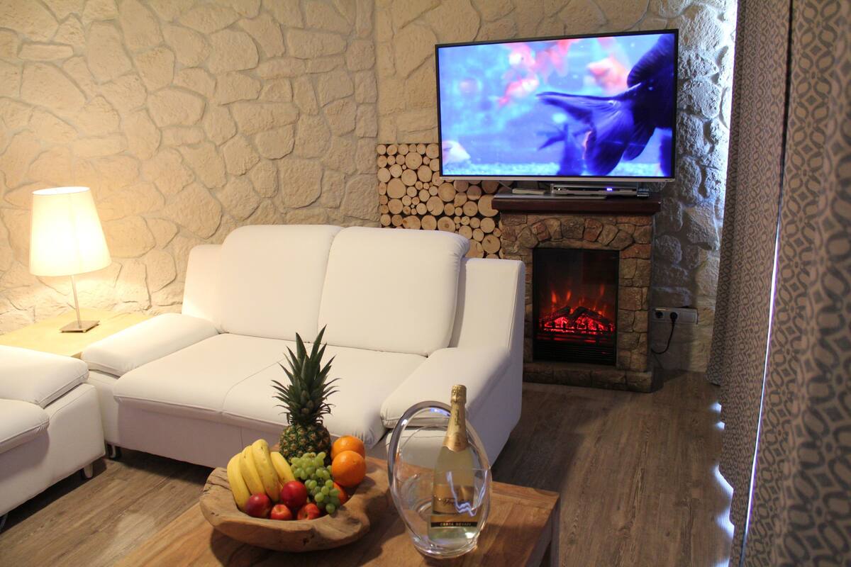 A living room with a white sofa, a wooden side table with a fruit bowl and a bottle, a television, and a stone wall with a fireplace.
