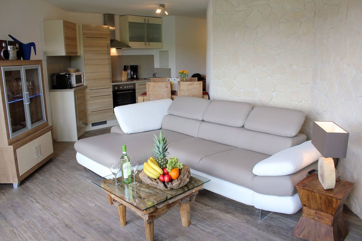 A modern living room with a beige sofa, a glass coffee table with fruit, and a kitchen in the background.