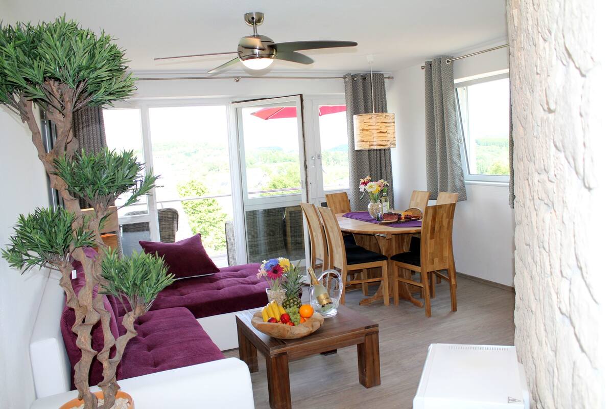 A living room with a ceiling fan, a dining area with a table and chairs, and a large window with curtains.