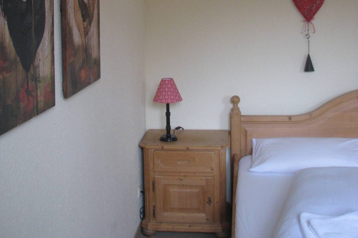 Une chambre à coucher avec une table de chevet en bois, une lampe, un lit et deux tableaux représentant des coqs, accrochés au mur.