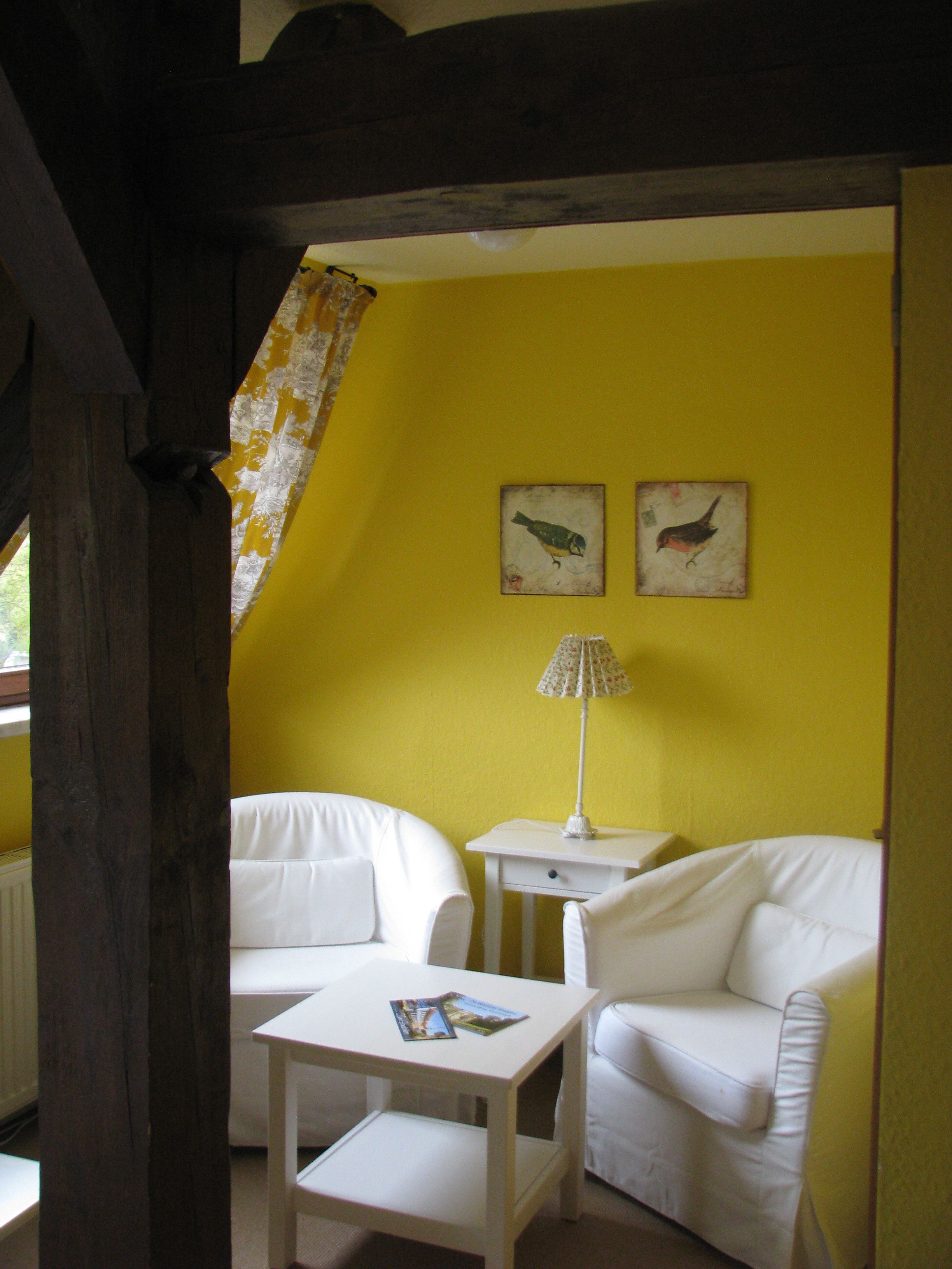 Een kamer met gele wanden, twee witte banken, een klein witte tafel en een lamp. Er hangen twee ingelijste foto's aan de muur.