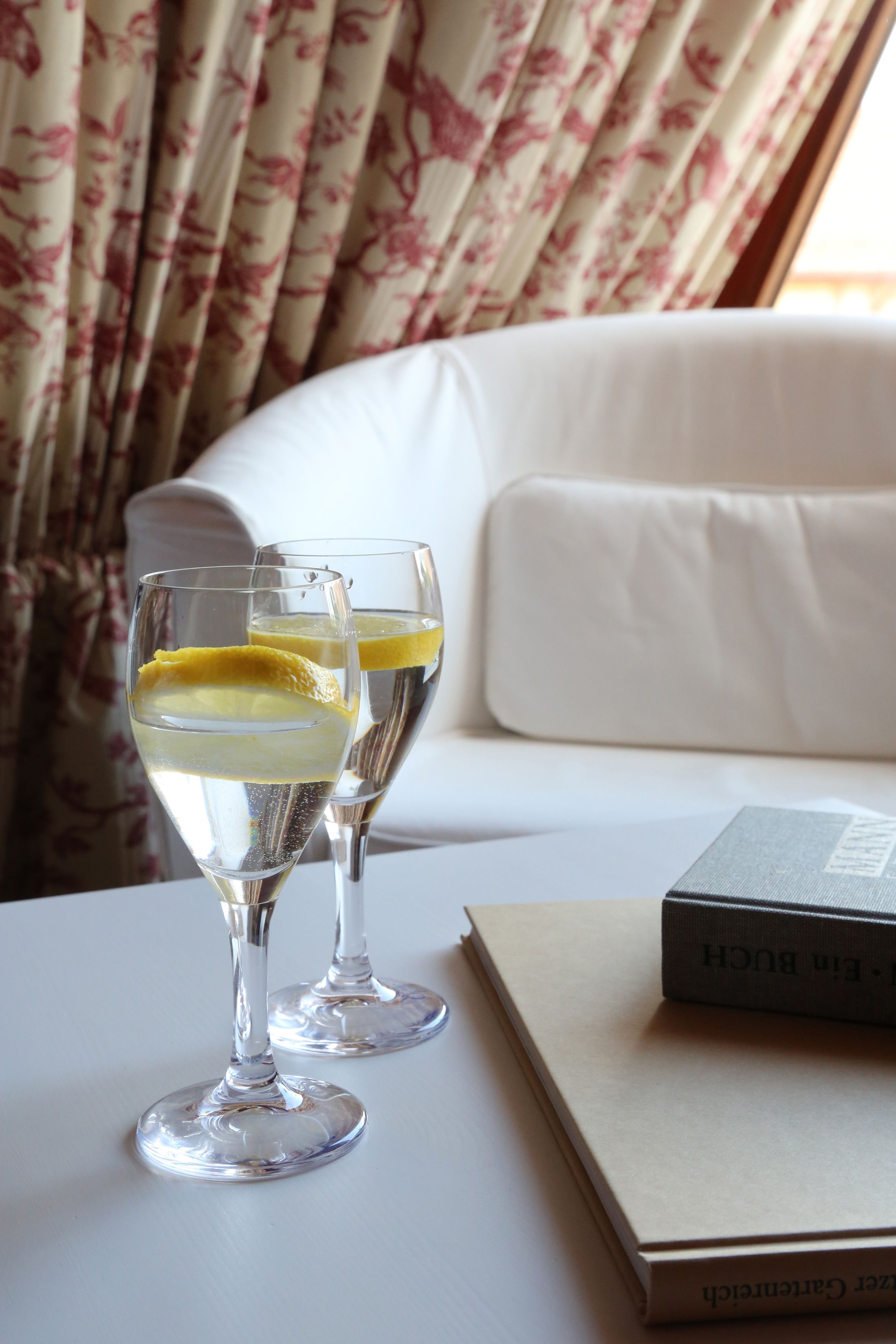 Two glasses of a clear beverage with a slice of lemon on a white table, a book titled "Ein Buch," and a white sofa with floral curtains in the background.