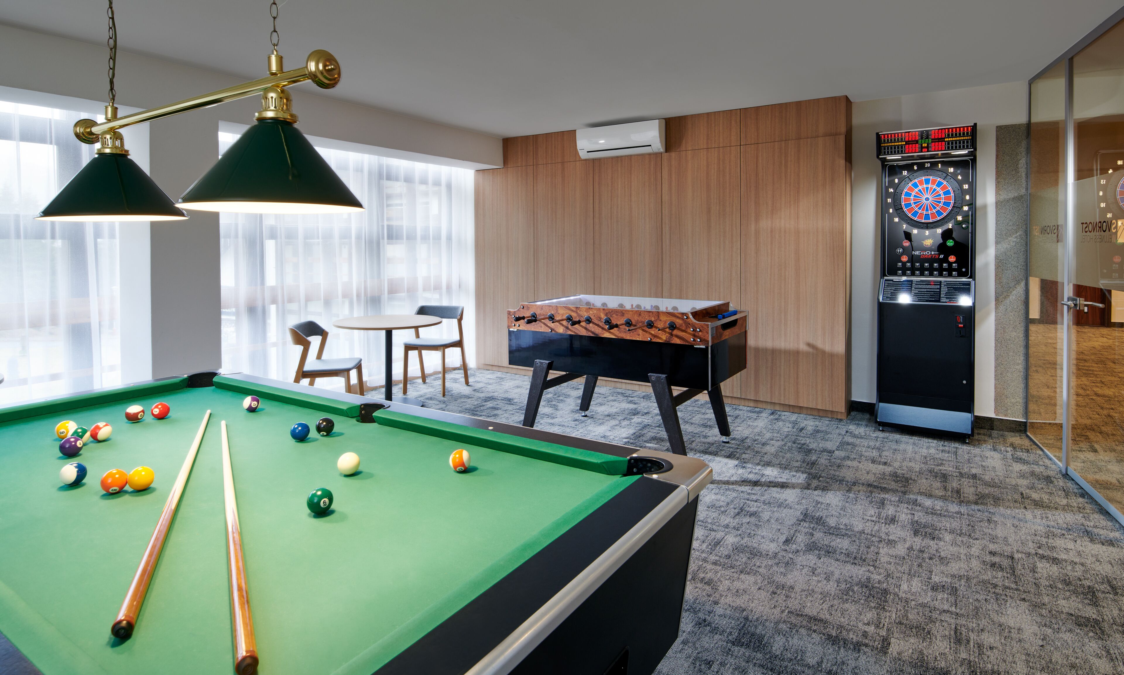 A pool table with balls, a foosball table, a dartboard, and a foosball table with a cloth cover.