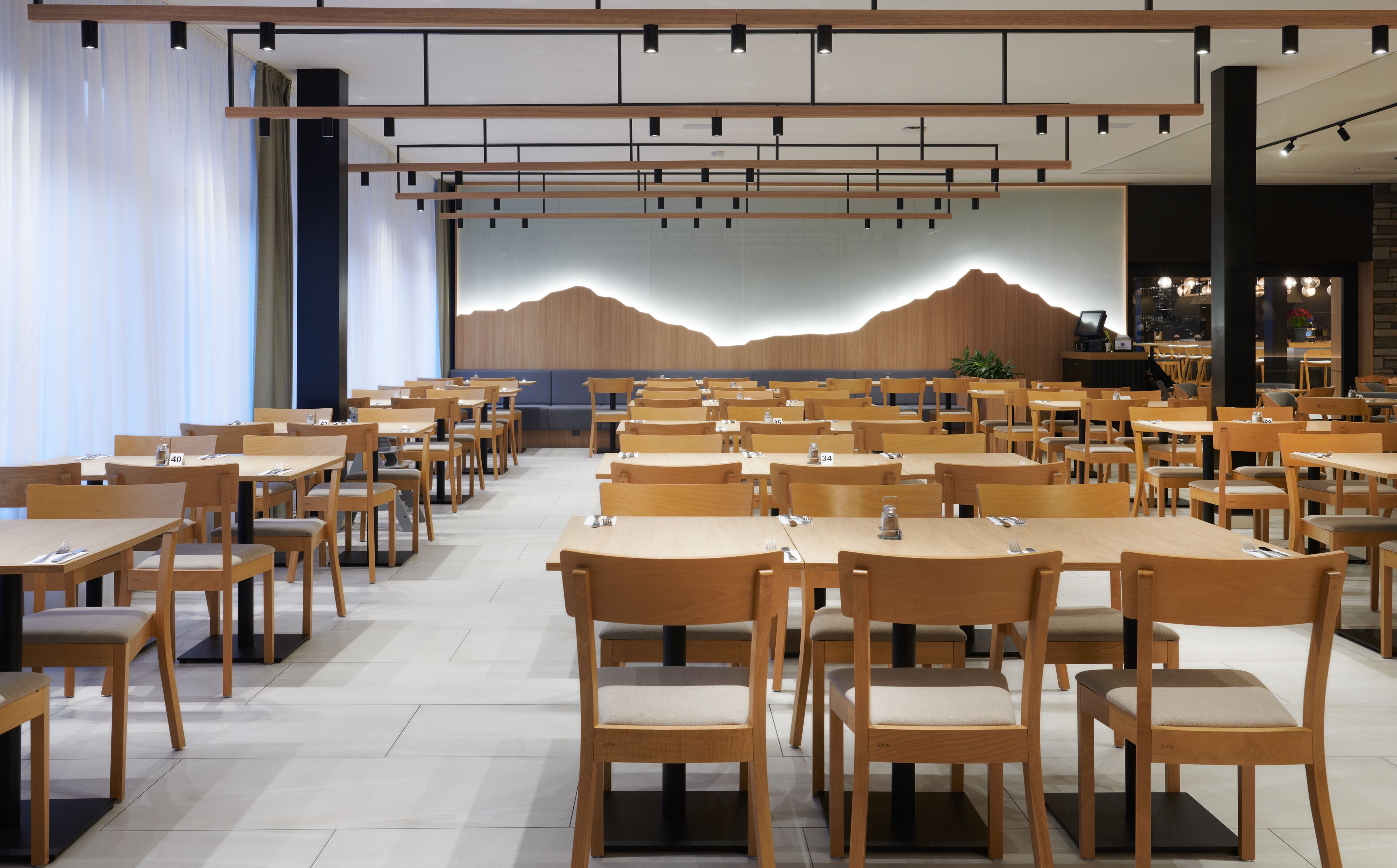 A spacious dining area with multiple wooden tables and chairs, a large wall mural, and a modern ceiling design.