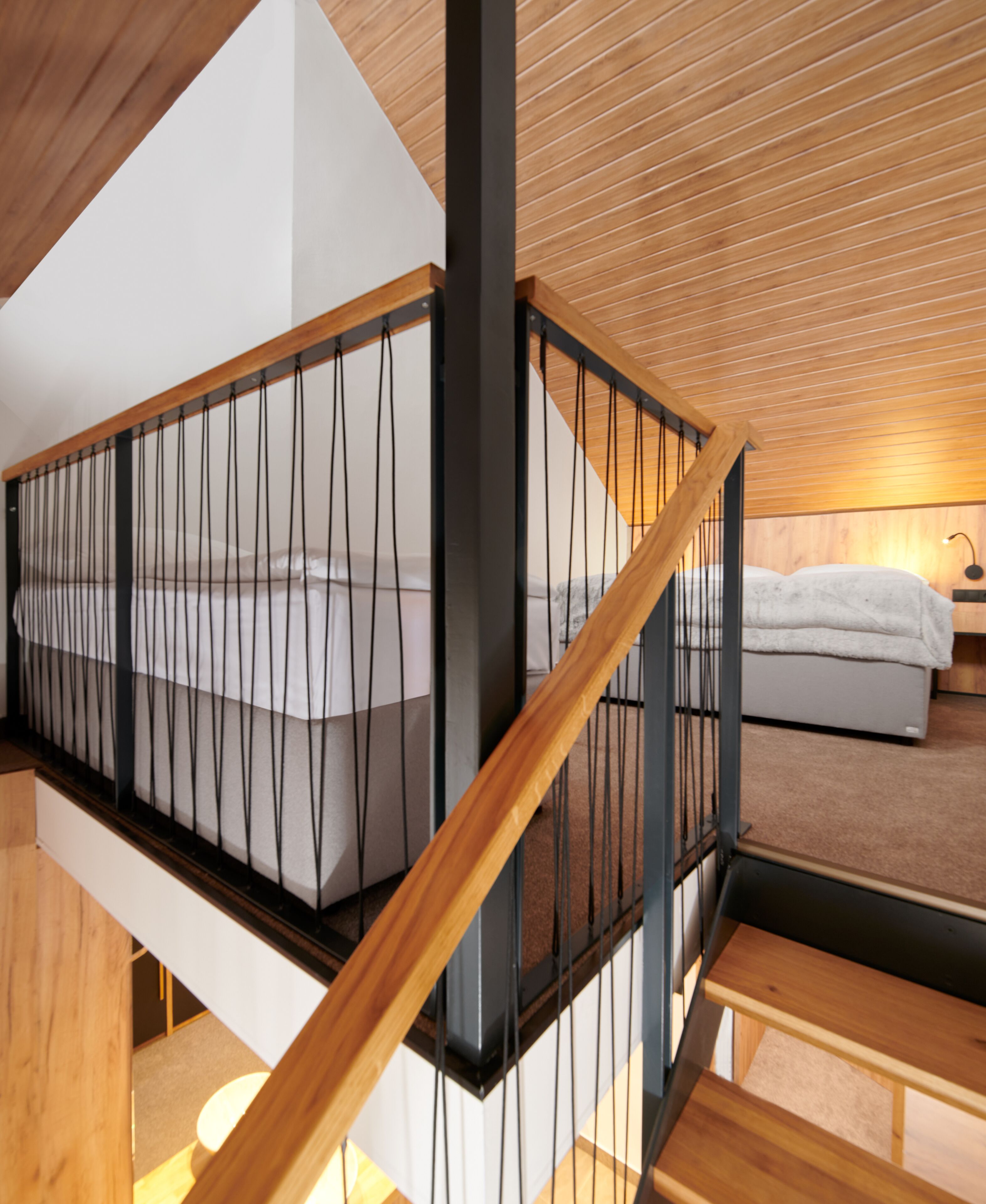 A modern staircase with a wooden handrail and black metal railings, leading to a bedroom with a bed and a nightstand.