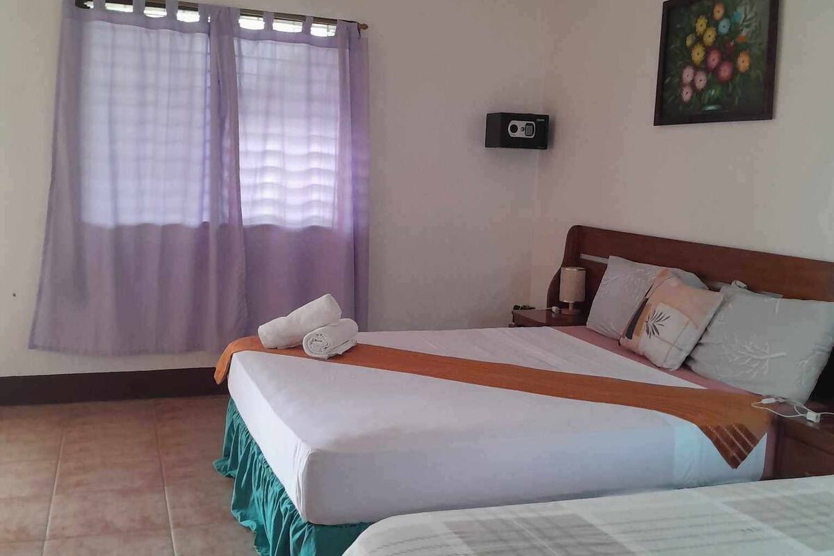 A hotel room with two beds, a ceiling fan, a window with curtains, a painting on the wall, and a small table with a lamp.