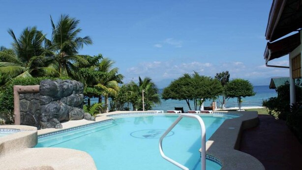 Une piscine bordée d’un mur de pierre et entourée de palmiers, avec des chaises longues et une vue sur l’océan.