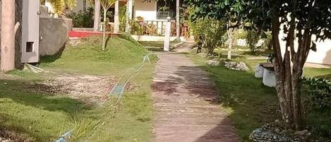 A paved pathway leading to a building with a red roof, surrounded by greenery and palm trees.