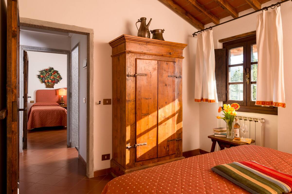 Une chambre avec une armoire en bois, un lit recouvert d’une couvre-lit à pois, une fenêtre avec des rideaux et une petite table sur laquelle est posé un vase de fleurs.