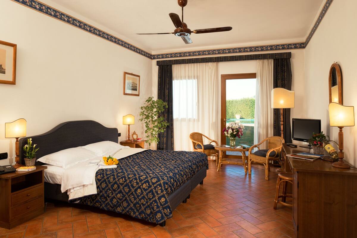 Une chambre d’hôtel avec un grand lit, deux tables de chevet, un bureau avec une chaise, une télévision et un ventilateur de plafond.