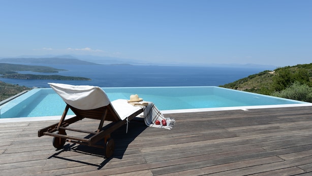 A wooden deck with a lounge chair and a small table, overlooking a clear blue pool and a scenic view of the sea and distant islands.