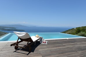 Une terrasse en bois avec un fauteuil de relaxation et une petite table, offrant une vue dégagée sur une piscine d’eau cristalline et un paysage pittoresque de la mer et des îles lointaines.