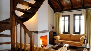 A cozy living room with a fireplace, striped sofa, wooden staircase, and large windows.