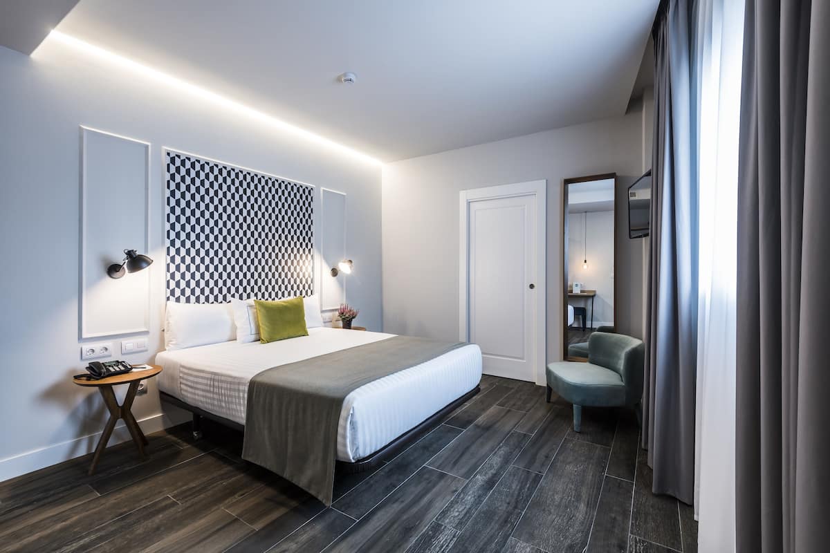 A modern hotel room with a large bed, a bedside table with a lamp, a patterned headboard, a chair, and a mirror.
