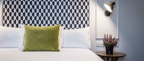 A neatly made bed with white and green pillows, a geometric patterned wall, and a wall-mounted lamp beside a small table with a potted plant.
