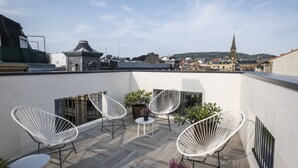 En tagterrasse med moderne hvide stole, et lille bord og plantekasser. En historisk bygning med et spiret tårn kan ses i baggrunden.