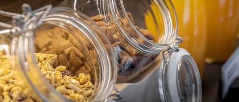 Una barra de desayuno con frascos de vidrio que contienen granola y otros productos para el desayuno, y una botella de jugo de naranja al fondo.
