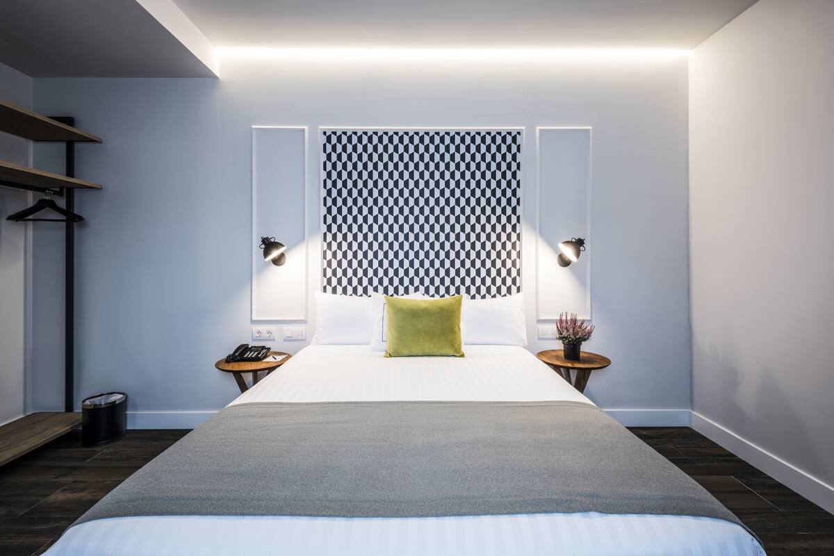 A modern hotel room with a large bed, bedside tables, a wall-mounted light panel, and a small side table with a plant.