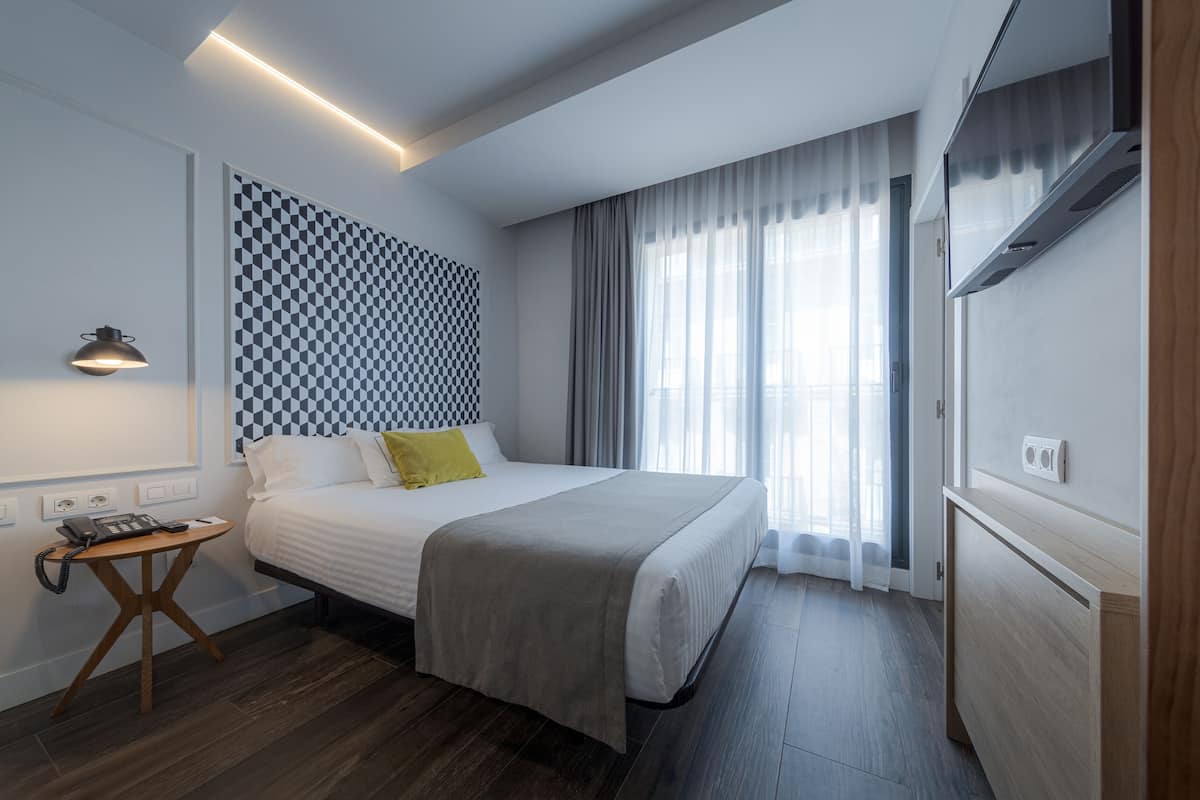 A modern hotel room with a large bed, a bedside table with a lamp, a patterned wall, and a large window with curtains.