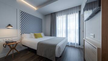 Una habitación de hotel moderna con una cama grande, una mesita de noche con lámpara, un papel tapiz estampado y un ventanal con cortinas.
