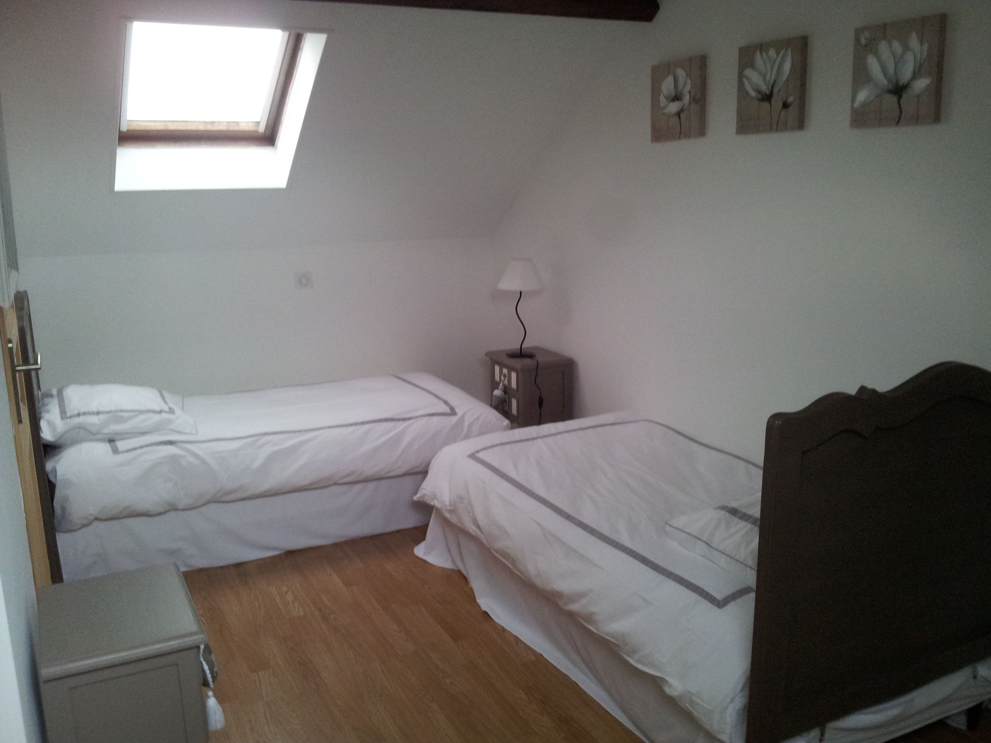 Ein Zimmer mit zwei Einzelbetten, einem kleinen Nachttischchen mit Lampe, Holzboden und einem Oberlicht.