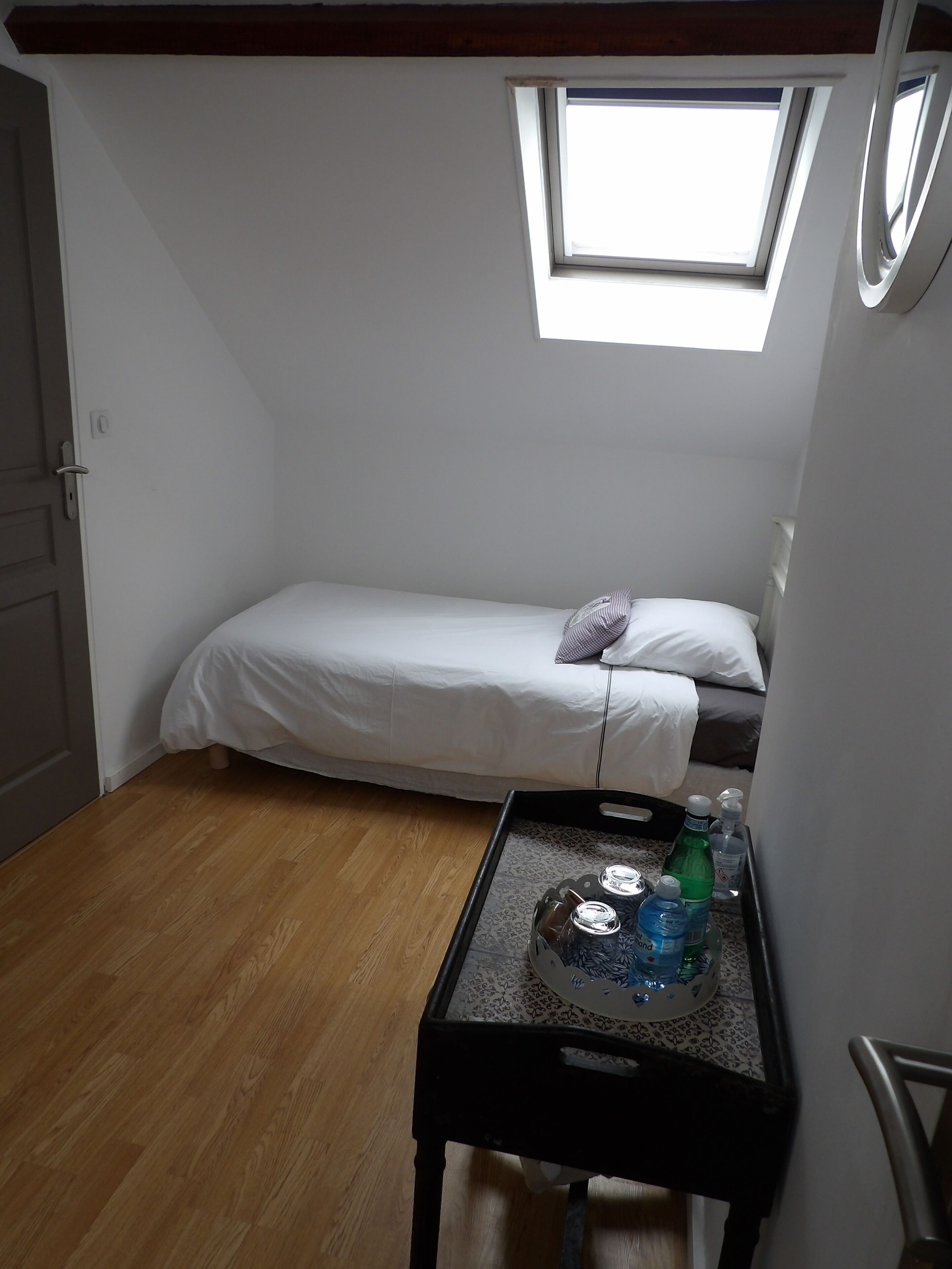 A small, single-bed room with a skylight, a wooden floor, and a small table with water bottles.