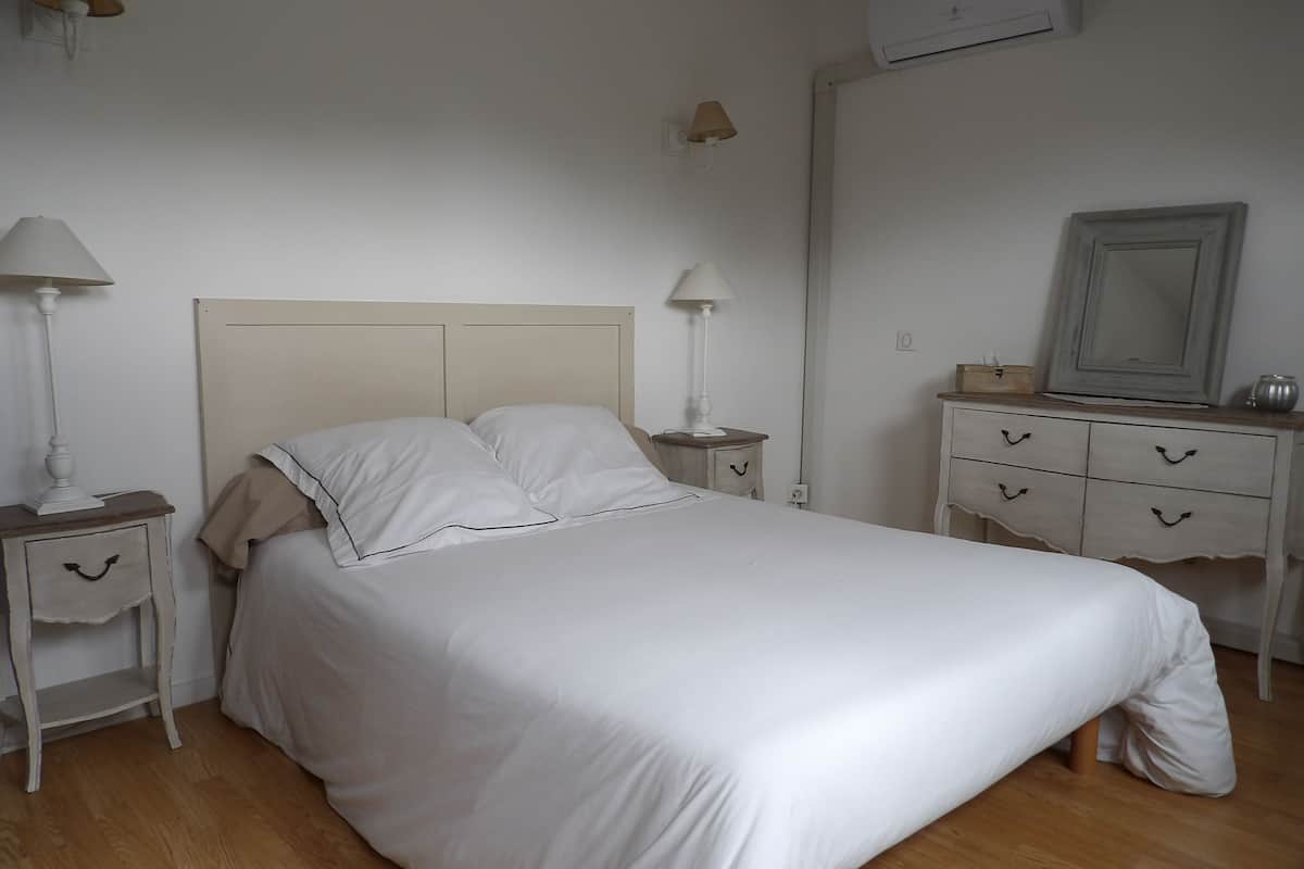 A neatly made bed in a room with a wooden floor, a dresser with drawers, a mirror, and two bedside lamps.