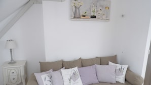 A cozy room with a beige sofa, a small white nightstand with a lamp, and a wall-mounted shelf with decorative items. - Le Clos Verlaine (Fampoux)
