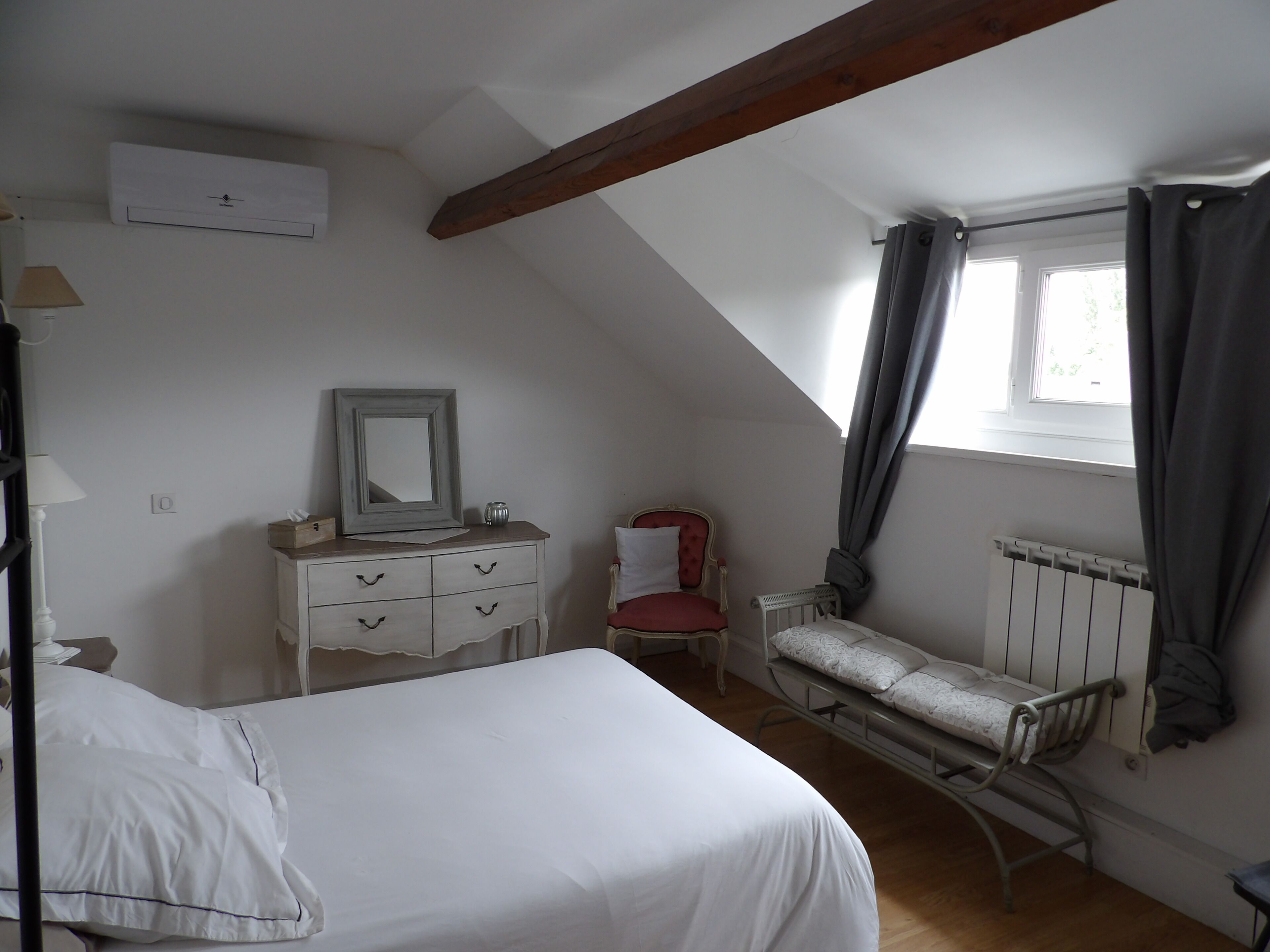 A bedroom with a bed, a dresser, a chair, a window with curtains, and a radiator.