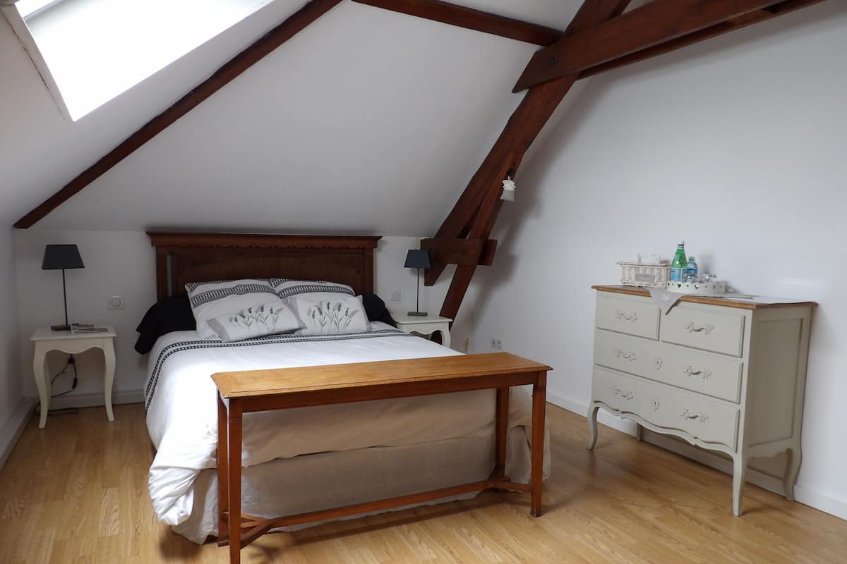 A bedroom with a sloped ceiling, wooden beams, a bed with patterned pillows, a bedside table with a lamp, a dresser, and a skylight.