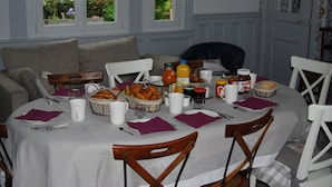 A dining table set with breakfast items, including bread, eggs, and beverages, in a room with a view of greenery through the windows. - Le Clos Verlaine (Fampoux)