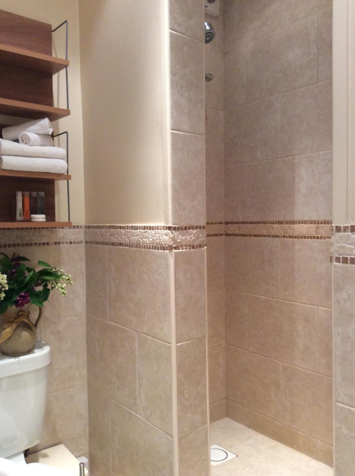 A shower area with beige tiles, a toilet, a shelf with towels, and a potted plant.