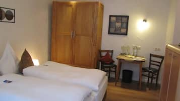A bedroom with two beds, a wooden wardrobe, a desk with a chair, and a small table with a lamp.